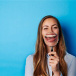 Brunette woman smiles and holds up a magnifying glass to her teeth for a post about fluoride, a natural mineral that prevents decay