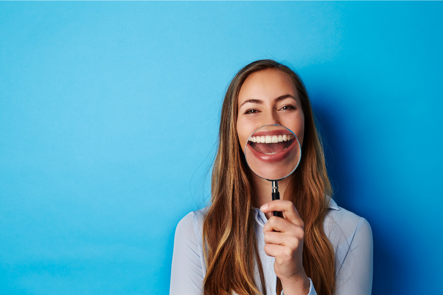 Brunette woman smiles and holds up a magnifying glass to her teeth for a post about fluoride, a natural mineral that prevents decay